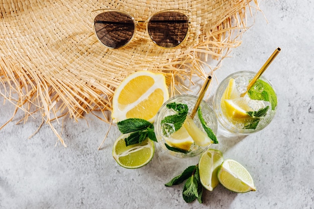 Cappello di paglia estivo e limonata di agrumi fresca in bicchieri e occhiali da sole su sfondo di marmo