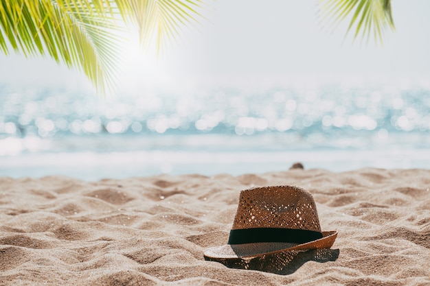 Cappello di paglia sulla vista sul mare tropicale della spiaggia della sabbia con la luce del bokeh della sfuocatura e della palma del mare calmo