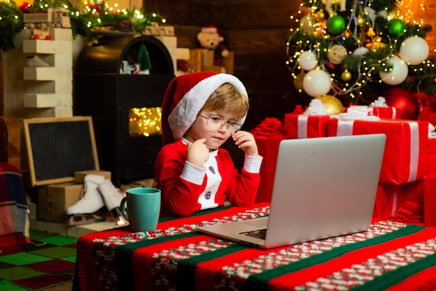 Cappello e costume della santa del ragazzino che si divertono. Acquista i regali di Natale online. Concetto di acquisto di Natale. Servizio regali. Piccolo genio. Piccolo aiutante di Babbo Natale. Bambino con laptop vicino all'albero di Natale