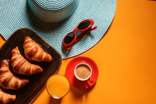 Cappello, occhiali da sole croissant con una tazza di caffè e un bicchiere di succo d'arancia su un tavolo giallo al sole