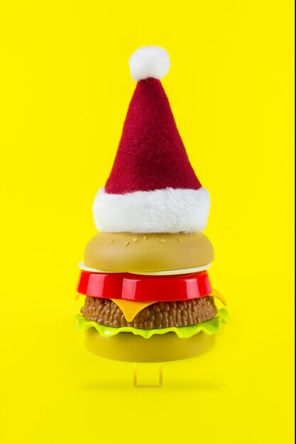 Cappello rosso-bianco della Santa sull'albero di natale di plastica dell'hamburger su fondo giallo. Hamburger di Natale