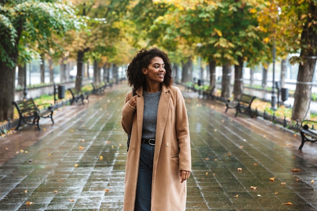 Cappotto da portare della bella giovane donna africana che cammina all'aperto al parco, portando lo zaino