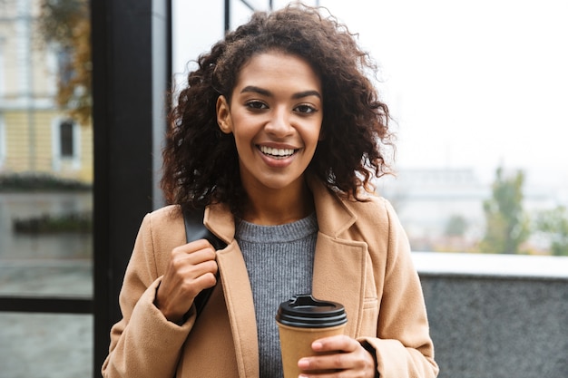 Cappotto da portare della giovane donna africana allegra che cammina all'aperto, che tiene tazza di caffè da asporto