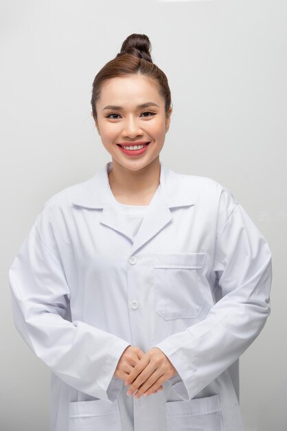 Cappotto da portare della giovane donna asiatica del medico che si leva in piedi sopra fondo bianco isolato con un sorriso felice e fresco sul fronte