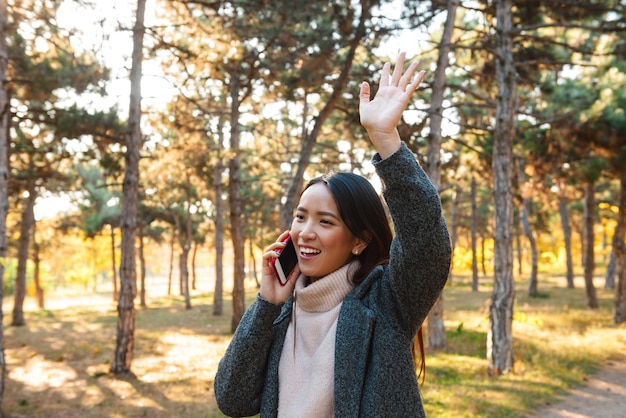 Cappotto da portare della giovane donna asiatica sorridente che cammina all'aperto al parco, parlando sul telefono cellulare