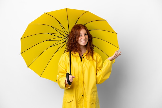 Cappotto e ombrello antipioggia della ragazza rossa dell'adolescente isolati su fondo bianco che allungano le mani al lato per invitare a venire