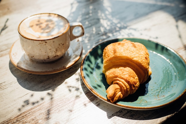 Cappuccino e croissant sono sul tavolo del bar. La luce del sole del mattino cade sul tavolo, appaiono belle ombre. Colazione deliziosa.