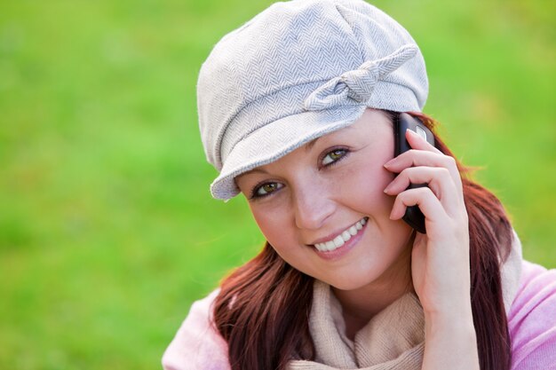 Cappuccio e sciarpa d&#39;uso sorridenti della giovane donna che parlano sul telefono sull&#39;erba