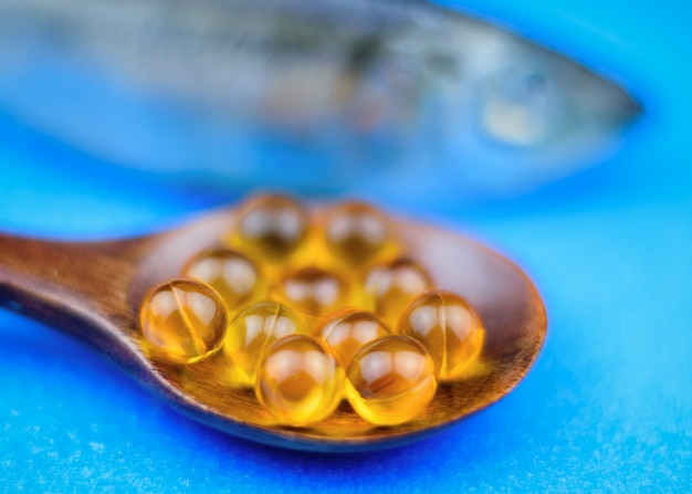 Capsule di olio di pesce in un cucchiaio di legno su uno sfondo blu con un pesce in background