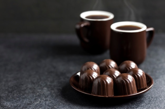 Caramelle di cioccolato su un piatto e due tazze di caffè caldo sulla superficie nera con lo spazio della copia