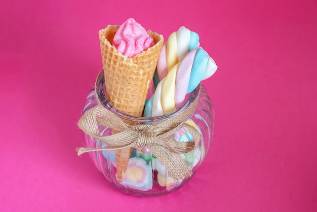 Caramelle e marshmallow con cono di cialda in vasetto di vetro su sfondo rosa Dolci caramelle di zucchero marshmallow e cono gelato Copyspace gratuito colori pastello