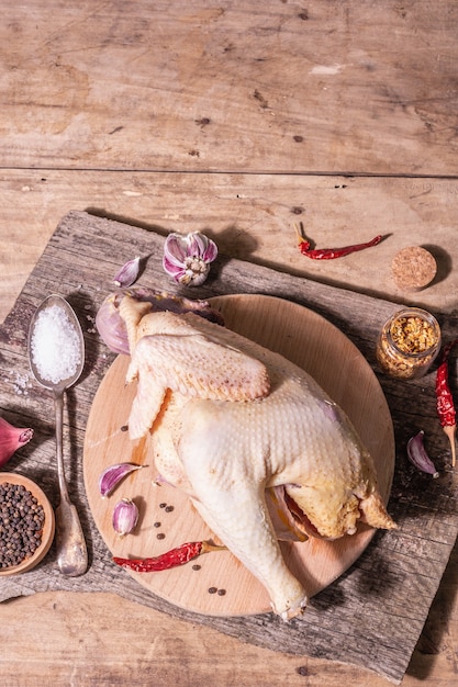 Carcassa di pollo intero crudo per uno stile di vita sano. Uccello da fattoria ruspante con cipolla rossa, spicchi d'aglio, spezie e sale marino. Una luce dura alla moda, un'ombra scura, un tavolo in legno vintage, vista dall'alto