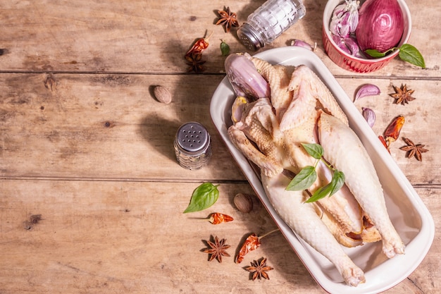 Carcassa di pollo intero crudo per uno stile di vita sano. Uccello da fattoria ruspante con spezie cinesi tradizionali in teglia da forno in ceramica. Una luce dura alla moda, un'ombra scura, un tavolo in legno vintage, vista dall'alto