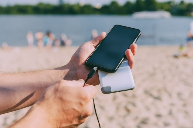 Caricatore portatile nelle mani di un uomo nello spazio della spiaggia. Powerbank ricarica il telefono.