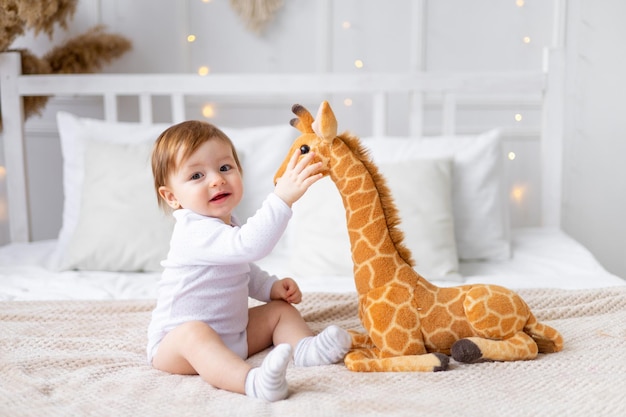 carina bambina che gioca con un giocattolo di giraffa di peluche seduta su un letto con una stanza luminosa dopo aver dormito e sorridendo