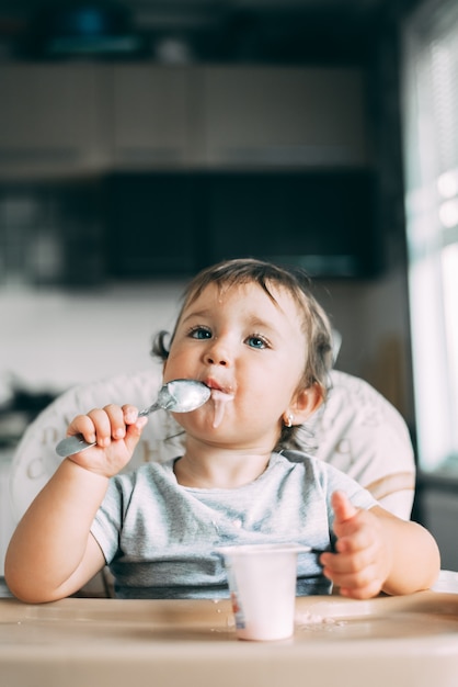 Carina, bambina che mangia yogurt in cucina durante il giorno in un seggiolone