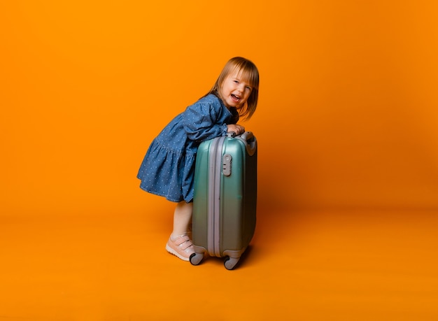 Carina bambina di 4 anni in un vestito di jeans blu con una valigia verde su sfondo giallo.