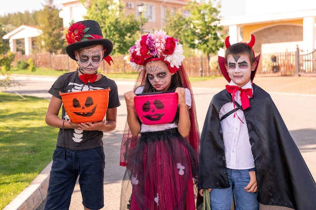 Carina bambina e due ragazzi con facce dipinte in piedi sulla strada contro i cottage e ti guardano mentre tengono le prelibatezze di halloween in cestini