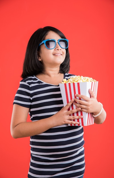 Carina bambina indiana che mangia popcorn mentre indossa occhiali da sole o occhiali 3D in un teatro. In piedi isolato su sfondo colorato