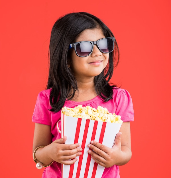 Carina bambina indiana che mangia popcorn mentre indossa occhiali da sole o occhiali 3D in un teatro. In piedi isolato su sfondo colorato