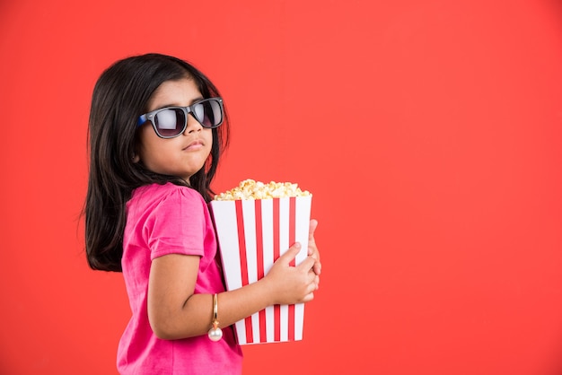 Carina bambina indiana che mangia popcorn mentre indossa occhiali da sole o occhiali 3D in un teatro. In piedi isolato su sfondo colorato