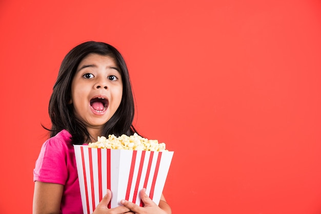 Carina bambina indiana che mangia popcorn mentre indossa occhiali da sole o occhiali 3D in un teatro. In piedi isolato su sfondo colorato