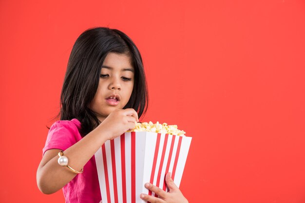 Carina bambina indiana che mangia popcorn mentre indossa occhiali da sole o occhiali 3D in un teatro. In piedi isolato su sfondo colorato