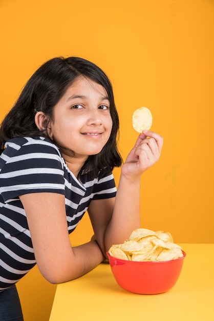 Carina bambina indiana o asiatica che mangia patatine o wafer di patate in una grande ciotola rossa, su sfondo giallo yellow