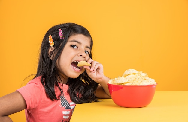 Carina bambina indiana o asiatica che mangia patatine o wafer di patate in una grande ciotola rossa, su sfondo giallo yellow