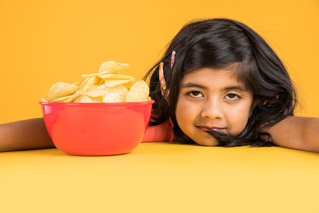 Carina bambina indiana o asiatica che mangia patatine o wafer di patate in una grande ciotola rossa, su sfondo giallo yellow