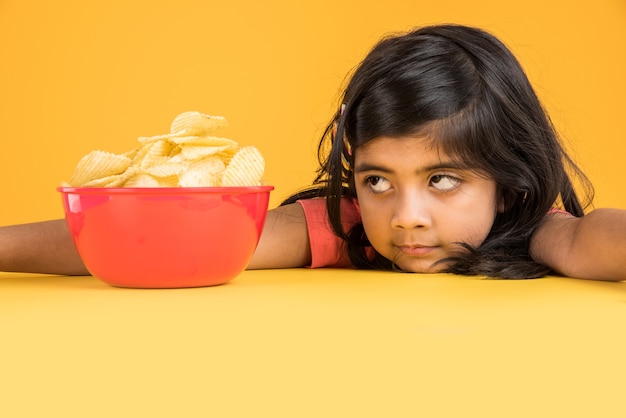 Carina bambina indiana o asiatica che mangia patatine o wafer di patate in una grande ciotola rossa, su sfondo giallo yellow