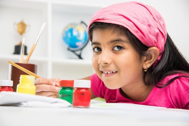 Carina bambina indiana o asiatica che si diverte a disegnare o dipingere con il pennello e dipingere su carta a casa