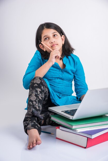 Carina bambina indiana o asiatica che studia sul laptop o lavora a un progetto scolastico mentre è sdraiata o seduta sul pavimento, isolata su sfondo bianco