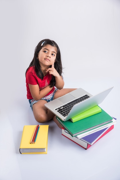 Carina bambina indiana o asiatica che studia sul laptop o lavora a un progetto scolastico mentre è sdraiata o seduta sul pavimento, isolata su sfondo bianco