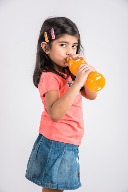 Carina bambina indiana o asiatica con una bottiglia di arancia o mango bevanda fredda o succo di frutta, bevendo o tenendo in piedi isolato su sfondo blu o bianco.