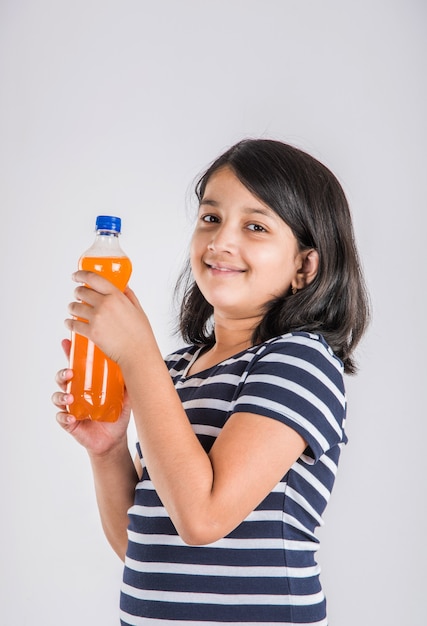 Carina bambina indiana o asiatica con una bottiglia di arancia o mango bevanda fredda o succo di frutta, bevendo o tenendo in piedi isolato su sfondo blu o bianco.