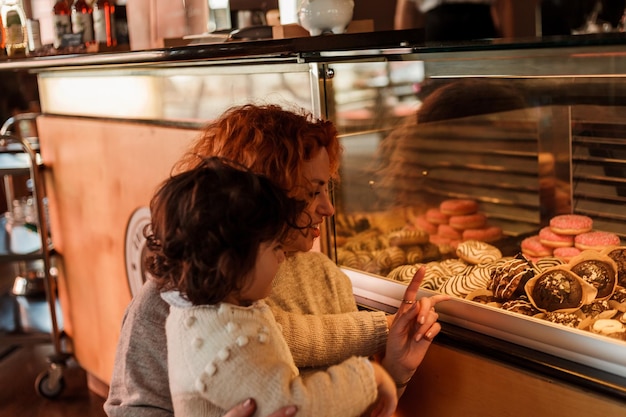 Carina bambina riccia di 3 anni con una giovane madre dai capelli rossi nella panetteria al bancone sceglie i pasticcini Cafe