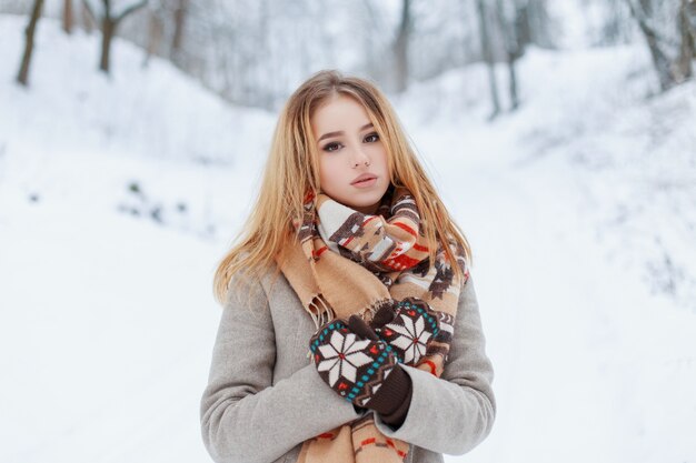 Carina bella giovane donna in un elegante cappotto invernale in guanti vintage di lana con una sciarpa di lana beige con un motivo multicolore che riposa nella natura in una fredda giornata invernale. Ragazza alla moda.