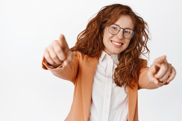 Carina commessa rossa che punta il dito alla telecamera sorridendo felice e scegliendo te congratulandosi con il vincitore invitando all'ufficio dell'azienda reclutamento hr in piedi su sfondo bianco