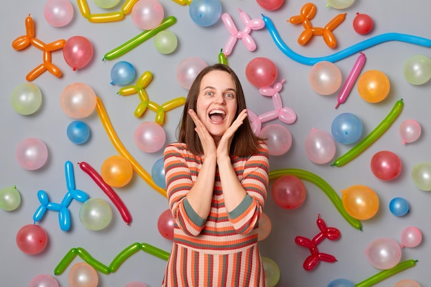 Carina donna affascinante sorridente che indossa un abito a righe in piedi contro il muro grigio con decorazione di palloncini esultando per la sua festa di compleanno a sorpresa che esprime emozioni festive positive