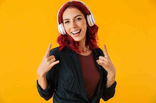 carina donna allegra con i capelli rossi ricci che ascolta musica con le cuffie e mostra il gesto delle corna isolato