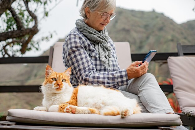 Carina donna anziana seduta all'aperto su una chaise longue in compagnia del suo bellissimo gatto Donna matura usa il cellulare mentre si rilassa in giardino