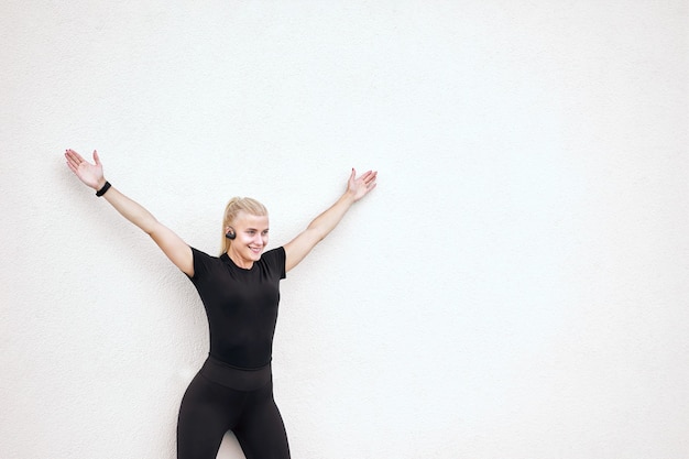 Carina donna esile allegra che indossa abiti sportivi neri, ascolta musica e stretching dopo l'allenamento.