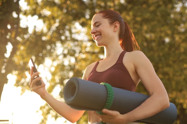 Carina donna sorridente che indossa tuta da ginnastica utilizzando smartphone e tenendo un tappetino da yoga al tramonto nel parco.