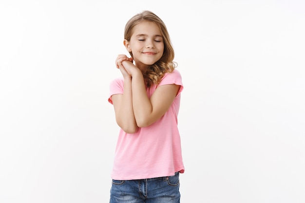 Carina e tenera ragazza adolescente femminile che sogna cose da ragazza, chiudere gli occhi premere i palmi sulla spalla scioccamente, sorridendo felice, immaginando vacanze fantastiche, immaginando un bel sogno, muro bianco