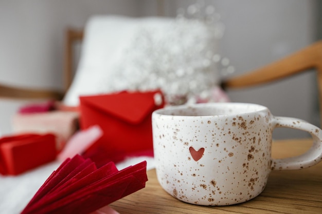 Carina ed elegante tazza di tè con cuore sul tavolo di legno contro la poltrona moderna con regali busta rossa e fiori bianchi Sorpresa mattutina di San Valentino per l'amato Buon San Valentino