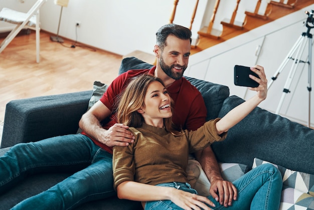 Carina giovane coppia in abbigliamento casual che si unisce e sorride mentre si fa selfie in casa