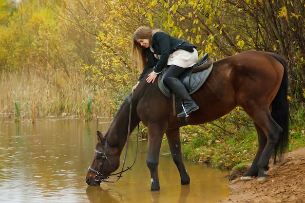 Carina giovane donna a cavallo nella foresta d'autunno in riva al lago. Il cavallo beve l'acqua nello stagno, la femmina accarezza la sua criniera. Cavaliere nel parco autunnale