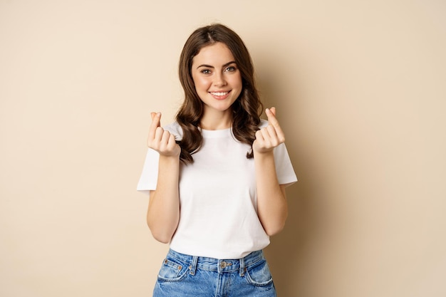 Carina giovane donna che mostra il segno dei cuori del dito e sorridente, esprime cura e amore, in piedi su sfondo beige.