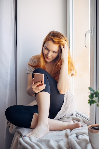 Carina giovane donna con lunghi capelli rossi utilizzando smartphone seduto sul davanzale della finestra a casa, foto verticale. Concetto di tecnologia e social network.